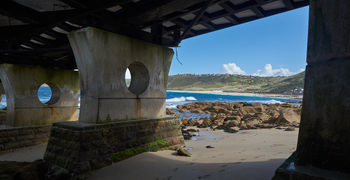 Sennen ramp This architectural photograph shows the Sennen ramp, an RNLI structure located on the coast at Sennen Cove in Cornwall, England, United Kingdom. Taken in the late morning during autumn, the image captures the ramp’s concrete supports with circular openings, providing a partial view of the adjacent sandy beach and rocky shoreline. In the background, houses are visible on the hillside and the sea displays waves breaking near the shore, characteristic of Sennen Cove’s coastal environment. The bright blue sky with scattered clouds indicates clear autumn weather. The composition highlights both the architecture of the RNLI ramp and the scenic coastal landscape of this location in Cornwall.